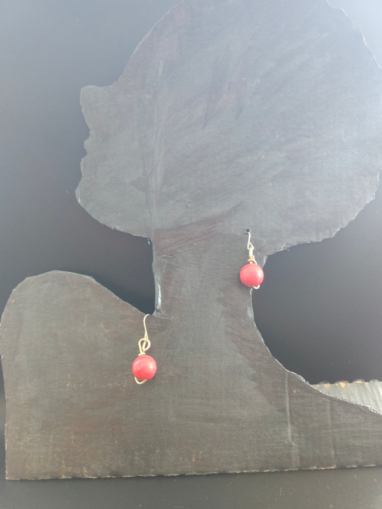 Tête mannequin portant des petites boucles d'oreilles rondes rouges. Elles sont entièrement modelées à la main et assemblées en France. Leurs perles en bois rouge apportent une touche de chaleur naturelle et d'authenticité à votre look. Le fil d'or (étain plaqué or 18 carats) qui entoure délicatement chaque perle crée un contraste élégant et raffiné. 