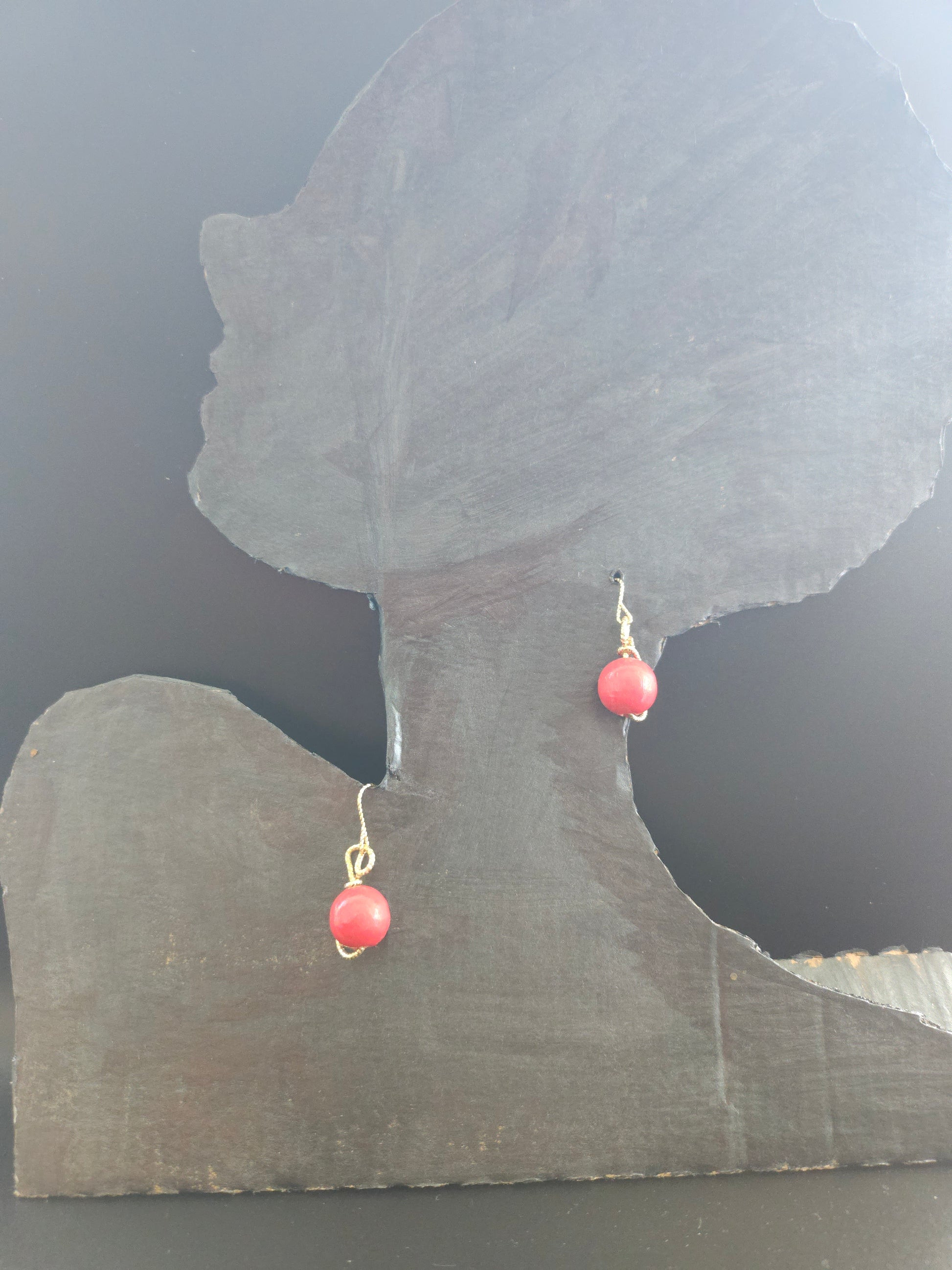 Tête mannequin portant des petites boucles d'oreilles rondes rouges. Elles sont entièrement modelées à la main et assemblées en France. Leurs perles en bois rouge apportent une touche de chaleur naturelle et d'authenticité à votre look. Le fil d'or (étain plaqué or 18 carats) qui entoure délicatement chaque perle crée un contraste élégant et raffiné. 